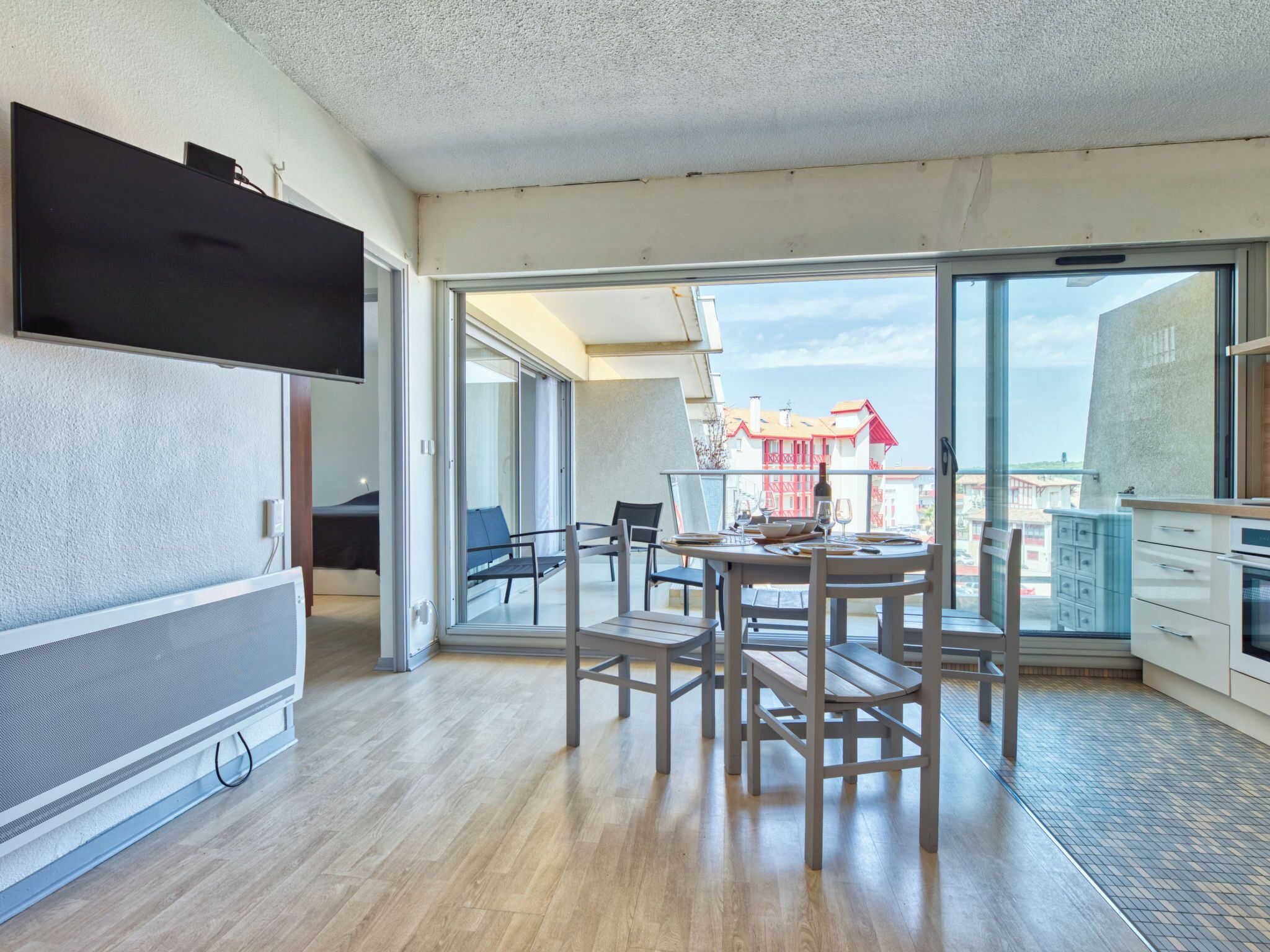 A modern apartment with a dining area, a TV, and a view of buildings through large glass doors.