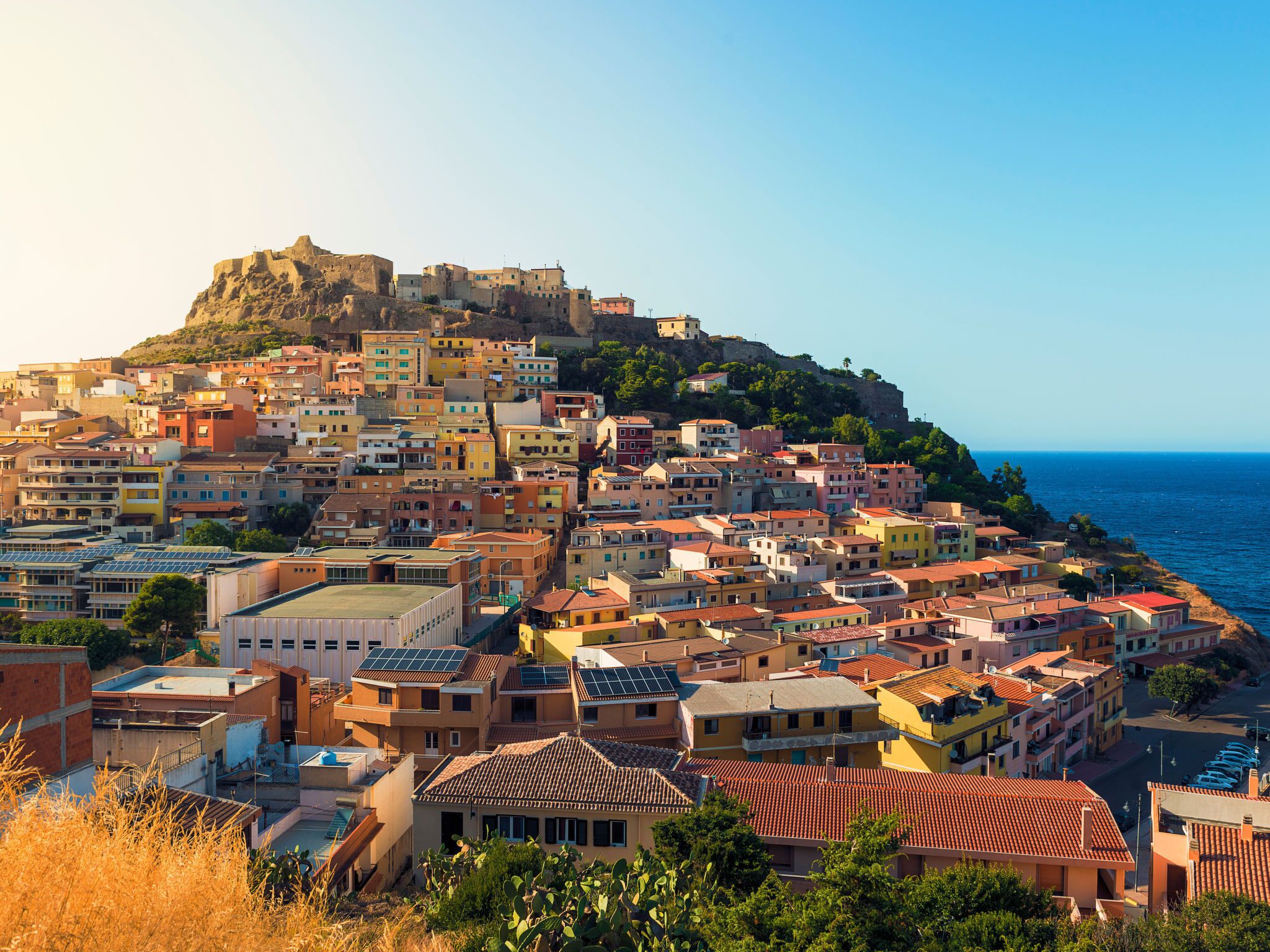 A coastal town with colorful buildings, a prominent hilltop fortress, and a clear view of the sea.
