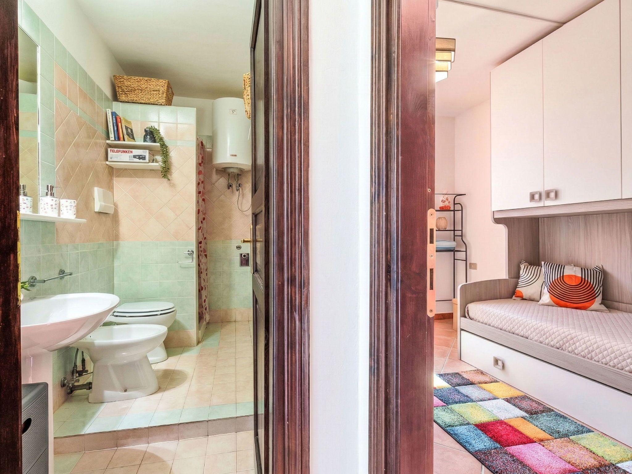 A modern bathroom with a sink, toilet, and shower. There is a colorful rug on the floor and a shelf with toiletries.