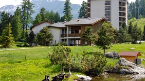 Un hotel con montañas al fondo, que presenta un edificio grande con balcones y una estructura más pequeña en primer plano.