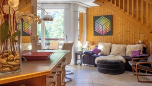 Una sala moderna con piso de madera, un sofá con cojines mullidos, una mesa de comedor con un jarrón de flores y un ventanal con vistas a la vegetación.