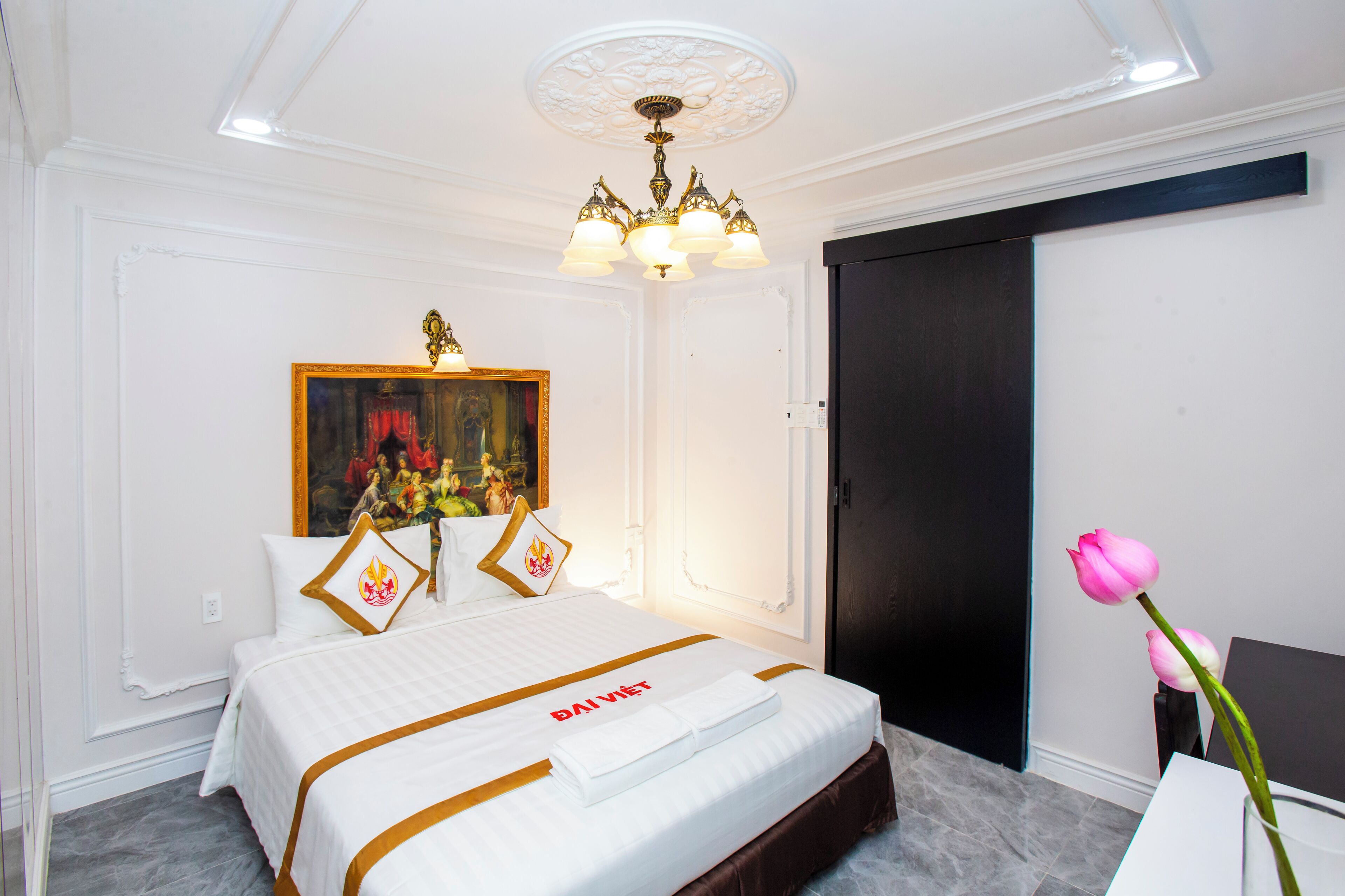 A hotel room with a bed, a painting, a chandelier, and a vase with a flower.