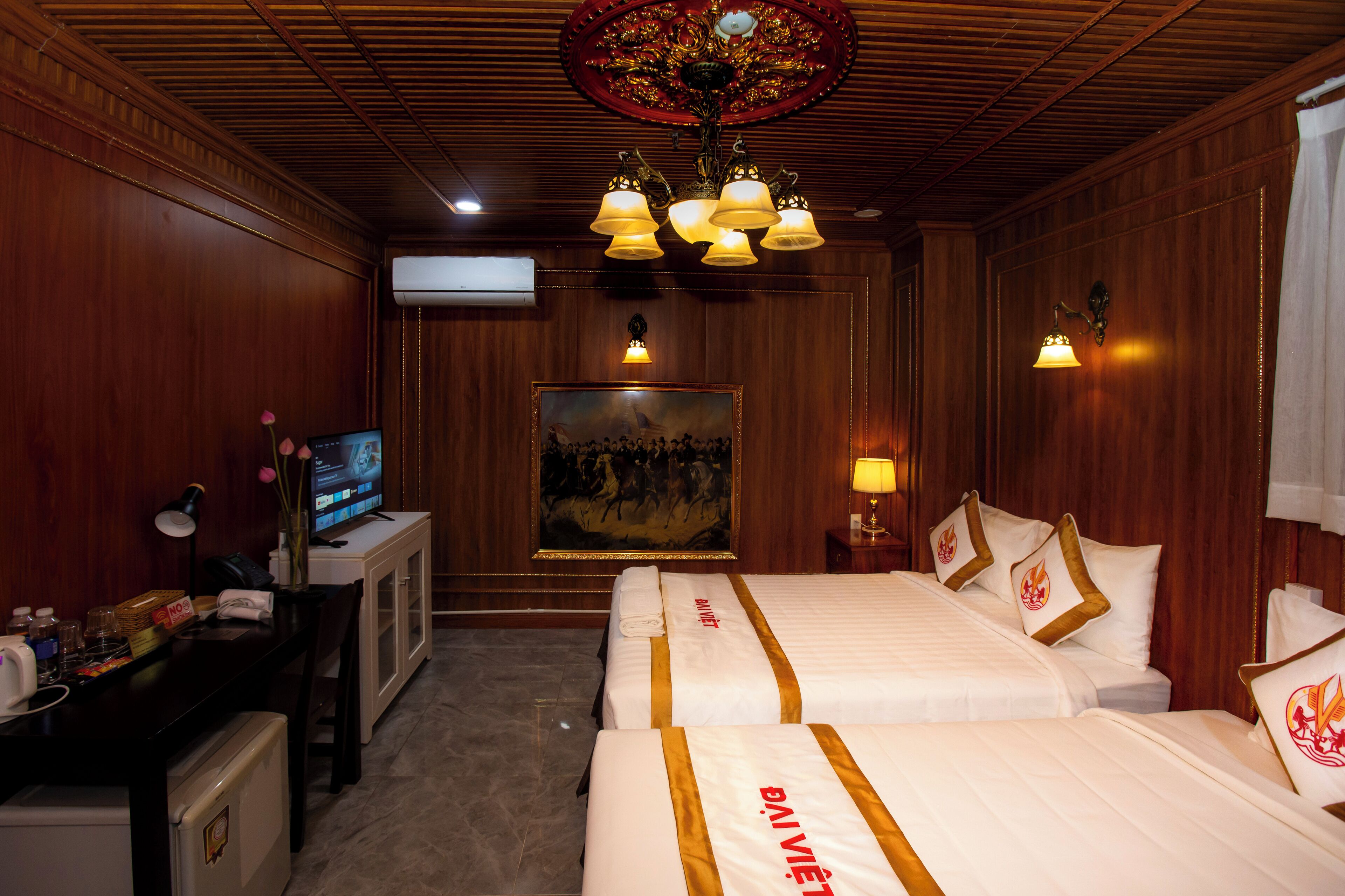 A hotel room with two beds, a desk with a television, a painting on the wall, and a chandelier.