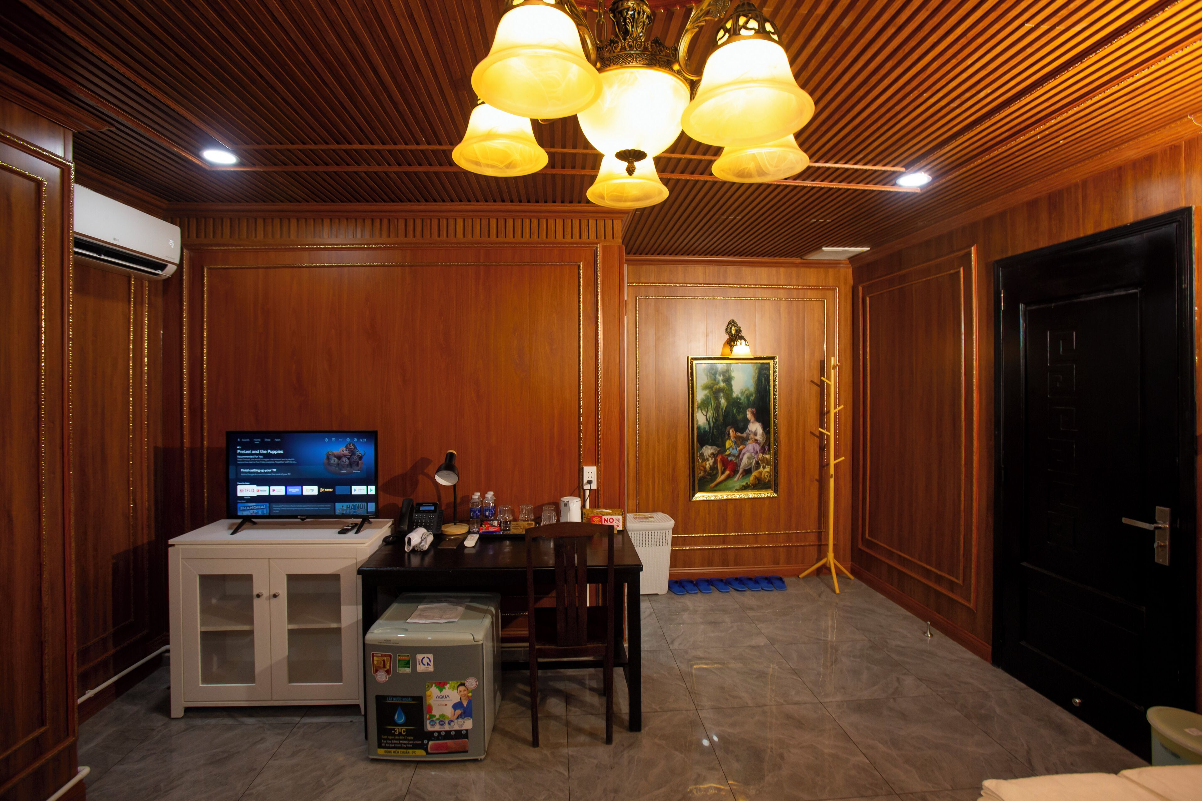 A room with wooden paneling, a desk with a computer, a chair, a small refrigerator, and a painting on the wall.