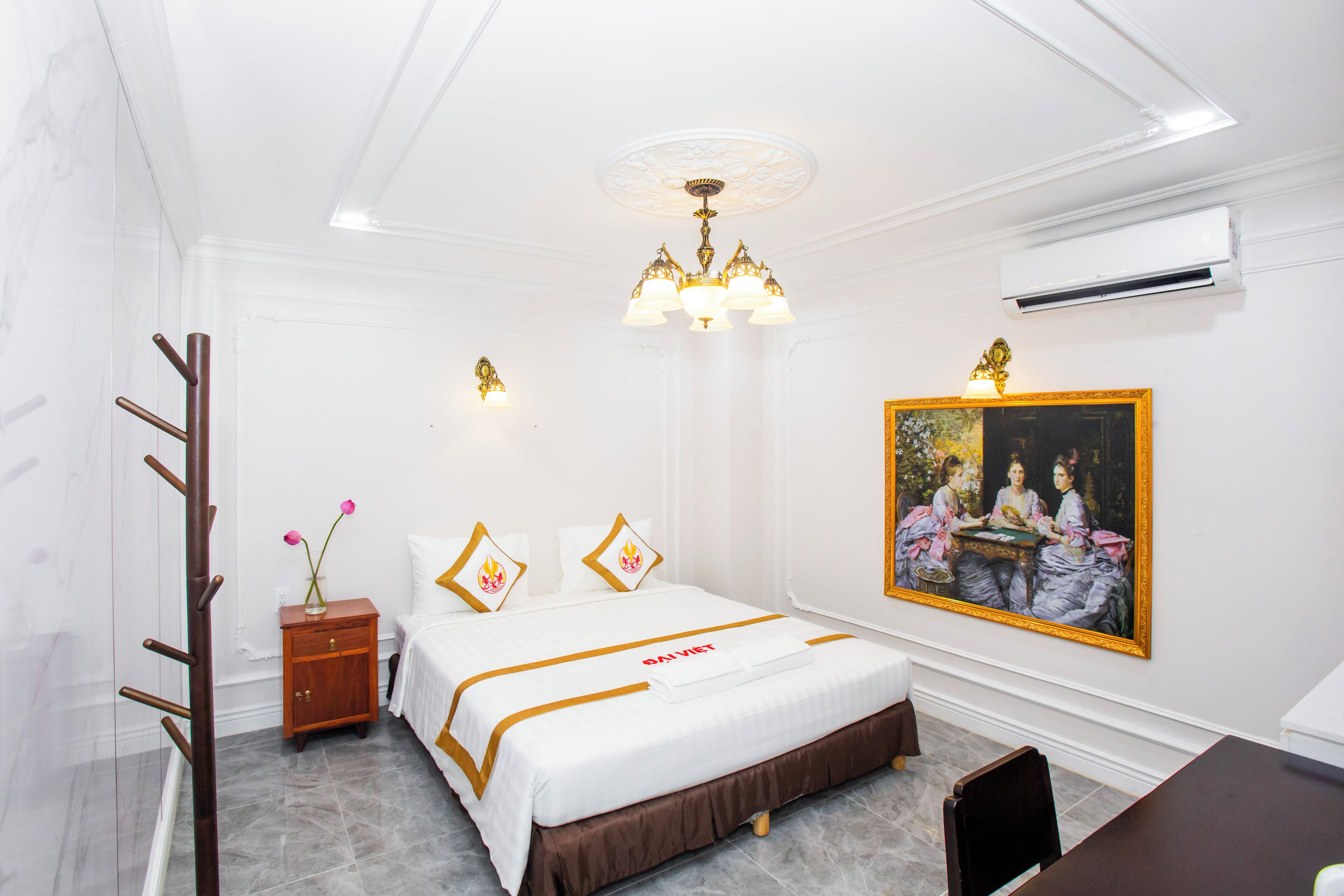 A hotel room with a bed, a nightstand, a desk, a chair, a painting on the wall, and an air conditioning unit.
