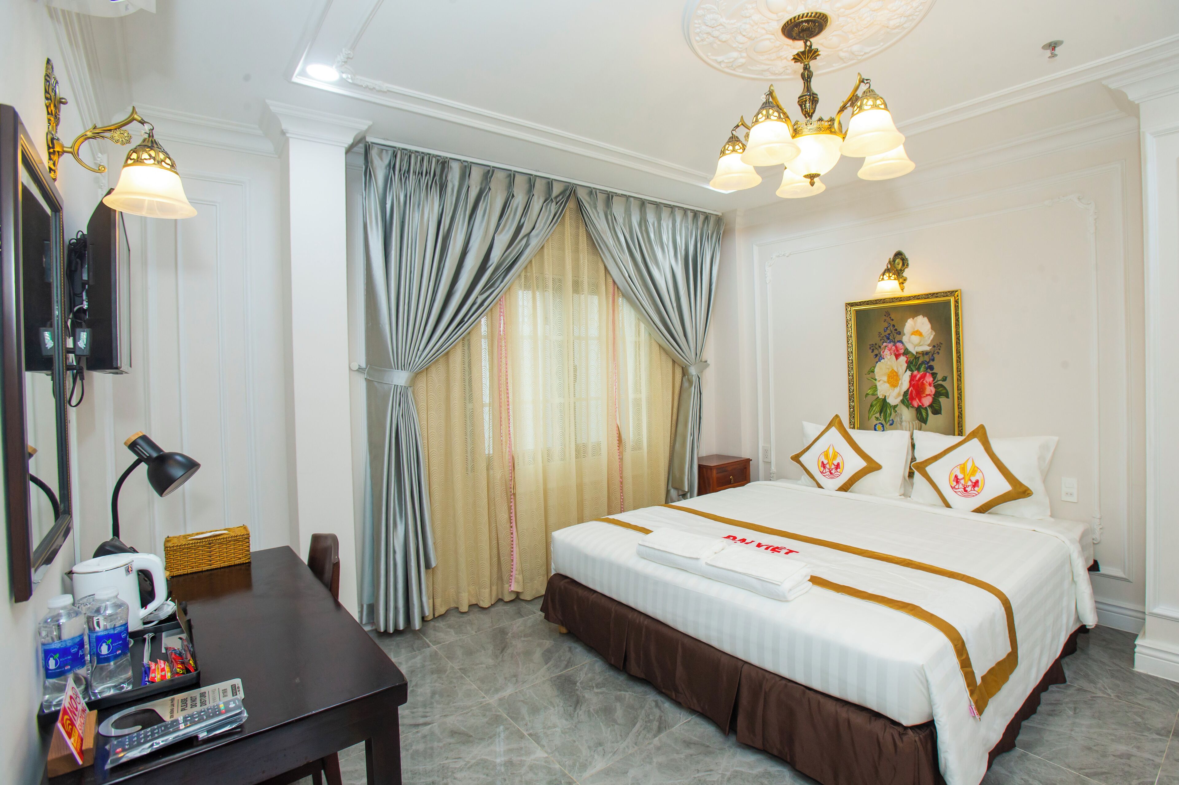 A hotel room with a large bed, a desk with a lamp, a television, and a painting on the wall.