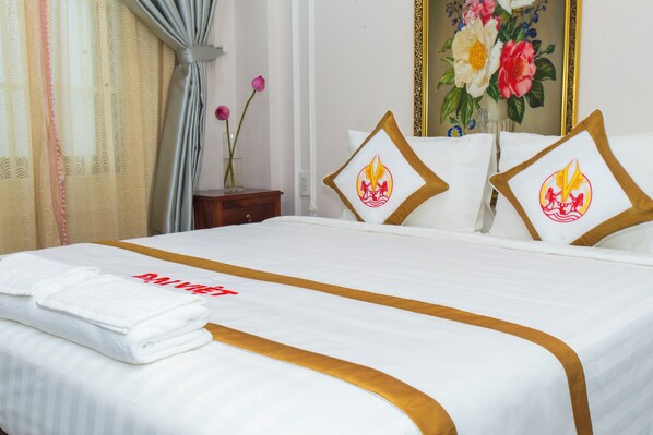 A neatly made bed with a white and gold bedspread, two pillows with a red emblem, and a folded white towel.