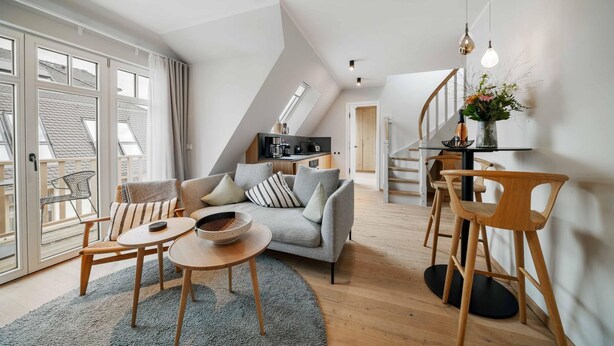 En moderne, åben loftslejlighed med en opholdsstue, hvor der er en grå sofa, træstole, et lille rundt bord og et køkken i baggrunden.