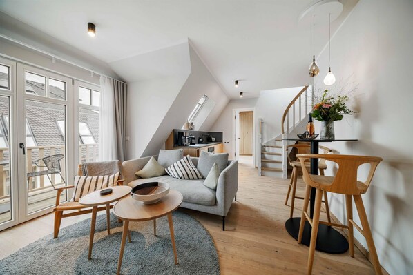 A modern loft apartment with a living area featuring a grey sofa, wooden chairs, a small round table, and a kitchen in the background.