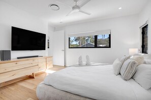 A modern bedroom with a large bed, a wooden dresser, a flat-screen TV, and a window with a view of trees and a house.
