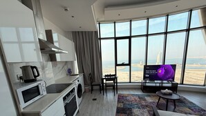 A modern kitchen with a microwave, kettle, and washing machine. There is a dining area with a table and chairs, and a TV displaying a purple animated character. The room has large windows offering a view of the city.