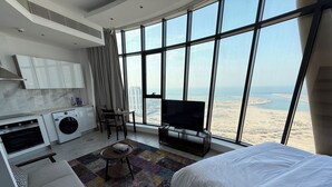 A modern hotel room with a large window, a bed, a TV, a small table, a sofa, a washing machine, and a kitchen area.