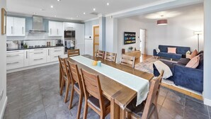 Una cocina moderna con armarios blancos, una zona de comedor con mesa y sillas de madera, y una sala de estar con un sofá azul y un televisor en la pared.