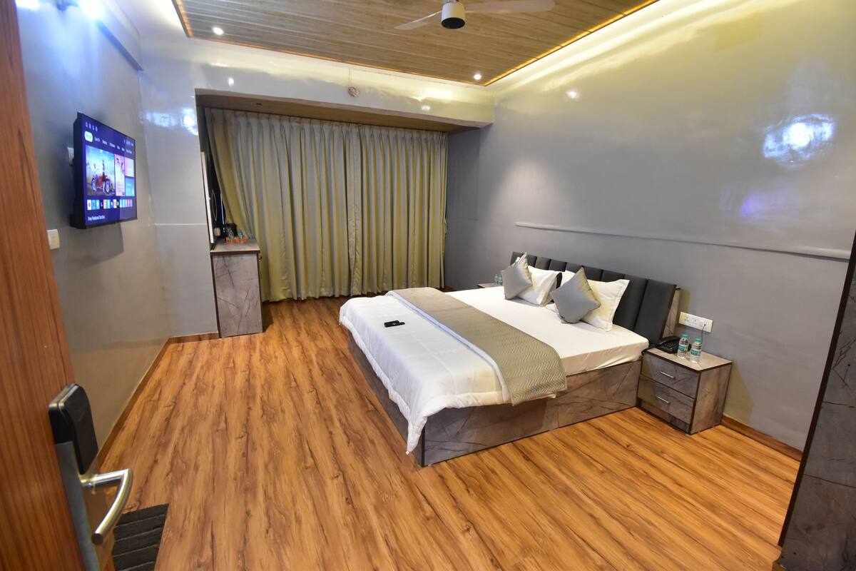 A modern hotel room with a wooden floor, a large bed with a headboard, a bedside table with a lamp, and a television mounted on the wall.