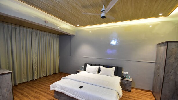 A modern bedroom with a wooden floor, a bed with white bedding, a ceiling fan, and a nightstand with a lamp and water bottles. - Hotel Evergreen  (Mumbai)