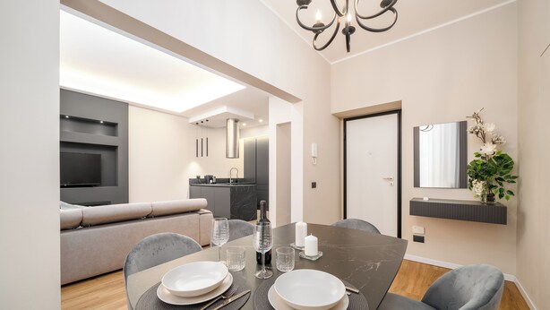 A modern dining area with a dark marble table set for two, surrounded by grey upholstered chairs. A sofa and a kitchen area are visible in the background.