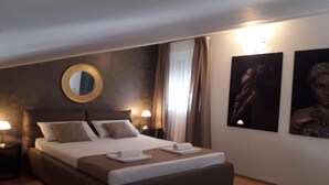 Hotellihuone, jossa on suuri sänky, kaksi yöpöytää valaisimineen, pyöreä peili seinällä ja kaksi kehystettyä kuvaa seinällä.