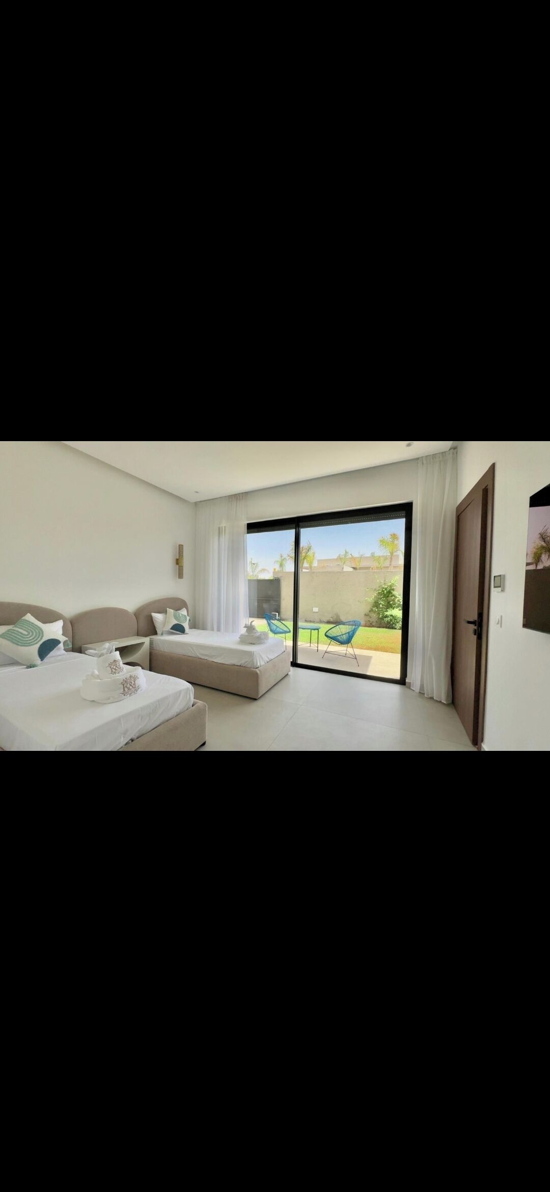 A hotel room with two beds, a large sliding glass door leading to a balcony, and a view of a garden.