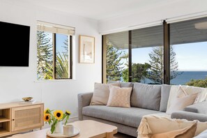 Una sala de estar moderna con un sofá gris, una mesita de madera, una mesa de centro y vista al mar a través de amplios ventanales.
