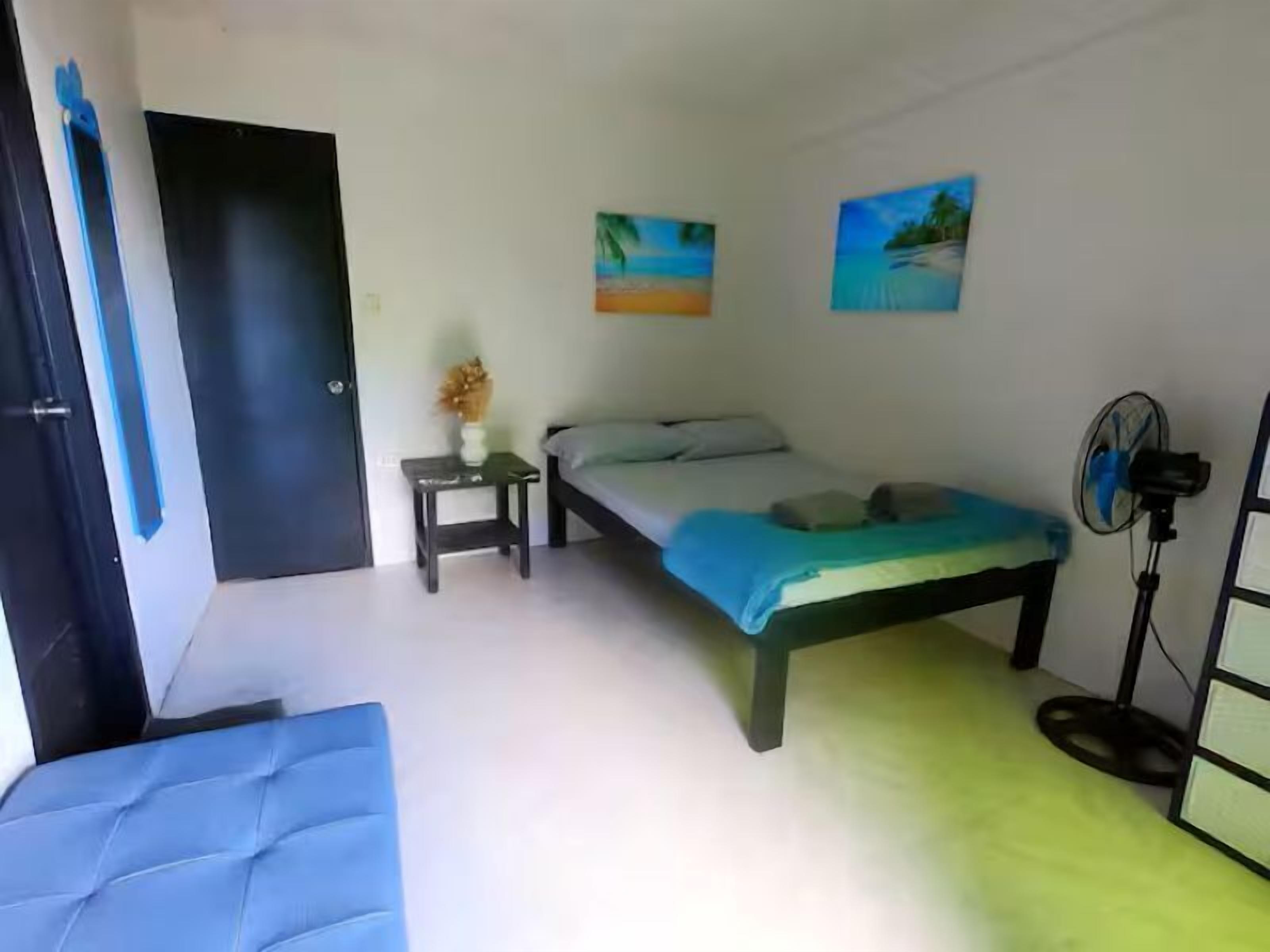 A bedroom with a bed, a small table with a vase, a standing fan, and two paintings on the wall.