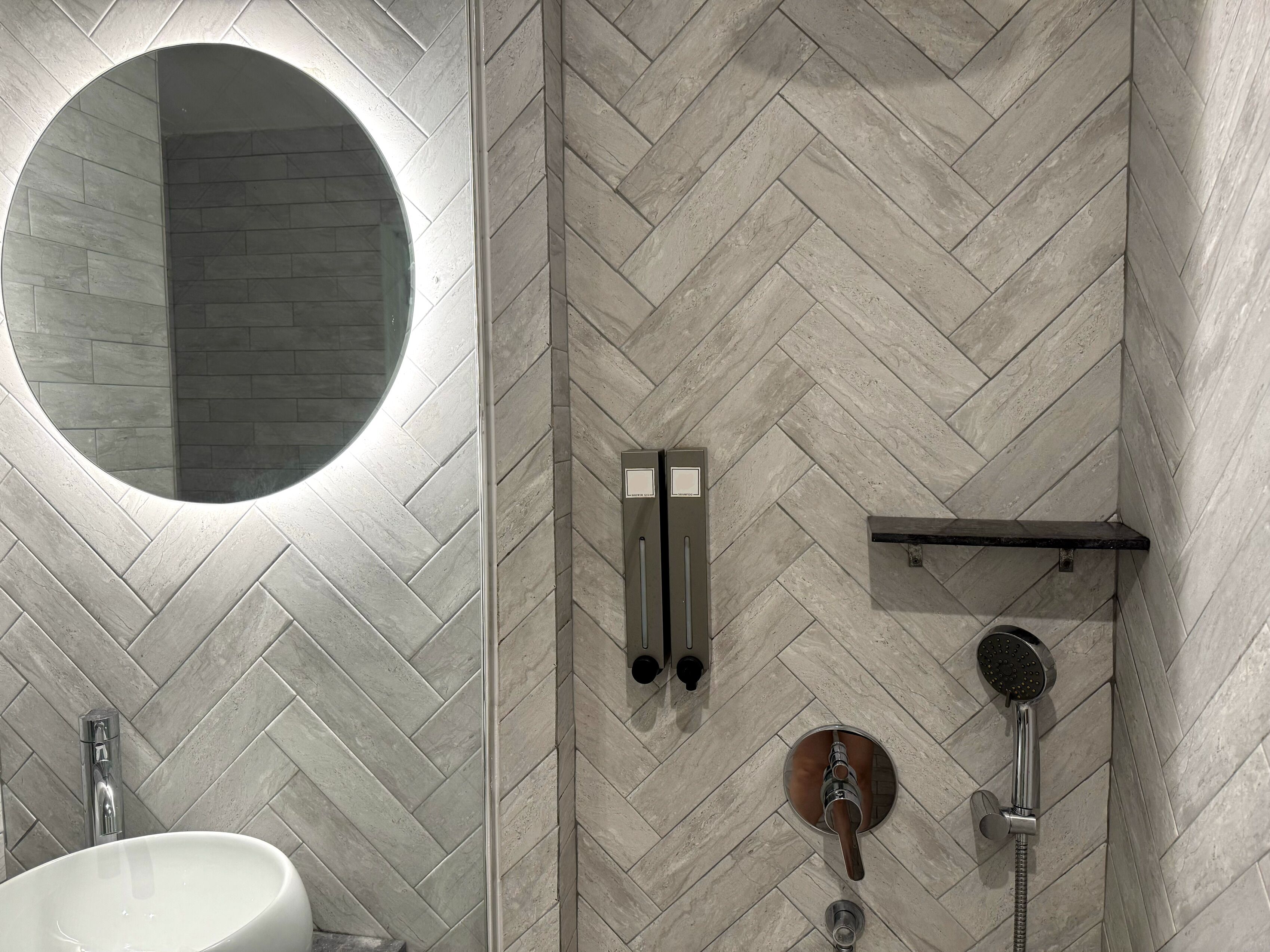 A modern bathroom with a herringbone tile pattern, a round mirror, a white sink, a towel rack, and a shower with a handheld showerhead.