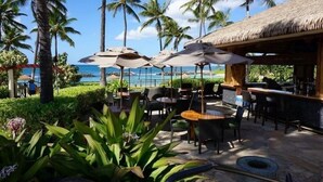 An outdoor seating area with umbrellas, tables, and chairs, overlooking a beach with palm trees and a thatched-roof structure. - Stunning Ocean View Luxury Suite - Ko Olina Beach Villas O421 End Unit (Kapolei)