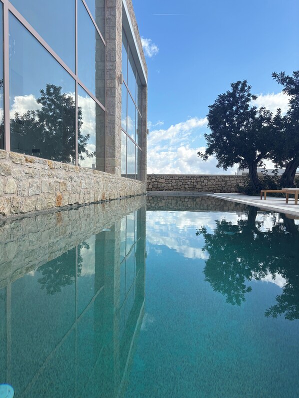 Piscina cristalina con paredes de piedra y ventanas amplias que reflejan árboles y cielo.