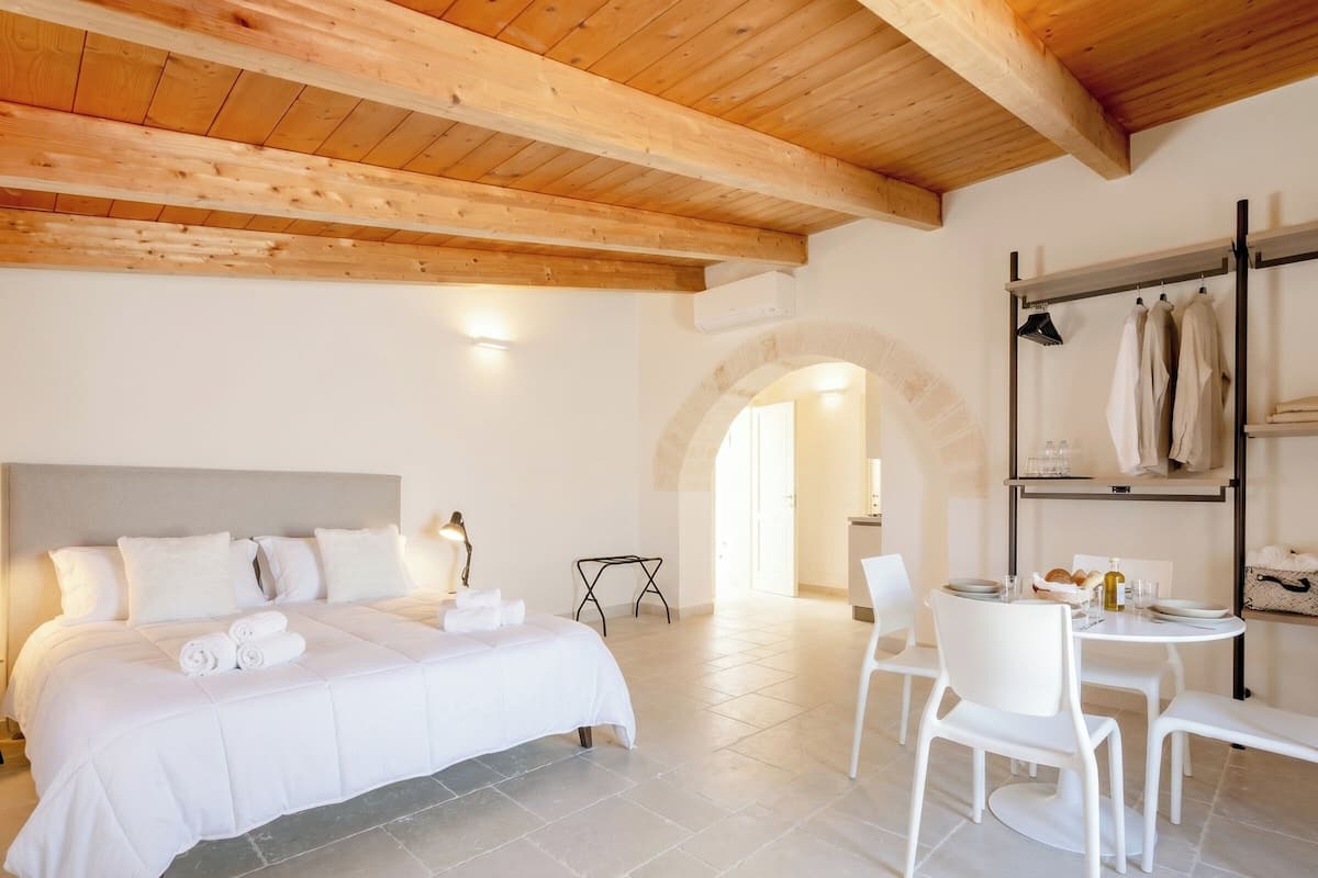 Une chambre d’hôtel avec un lit, une table et des chaises, une étagère avec des serviettes et une porte en arc de cercle.