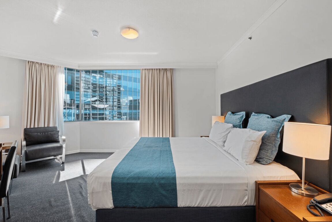 A hotel room with a large bed, a desk, a chair, and a view of the city through large windows.