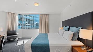 A hotel room with a large bed, a desk, a chair, and a view of the city through large windows.