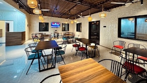 A modern dining area with multiple tables and chairs, a television screen, and a large window providing a view of the outdoors. - Hotel Vanvira (Mawal)