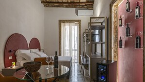 A hotel room with a bed, a dining table with chairs, a glass-top table with a bowl and wine glasses, a minibar, and a wall with wine bottles displayed. - Ballarooms Experience (Palermo)