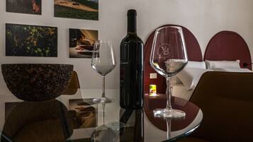 A hotel room with a glass table, wine bottle, and wine glasses. There are framed pictures on the wall and a bed with a red headboard.