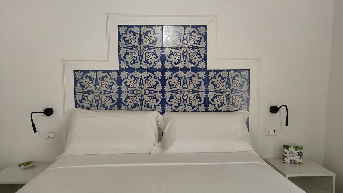 A bedroom with a white bed, two white pillows, a decorative blue and white headboard, and a small bedside table with a book.