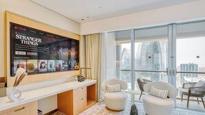 A modern hotel room with a large window, a flat-screen TV displaying a Netflix show, a desk with a laptop, and two comfortable chairs.