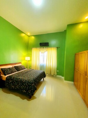 A bedroom with green walls, a bed with a floral bedspread, a nightstand, a lamp, a window with curtains, and a wardrobe.