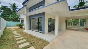 A modern two-story house with a large glass door and windows, a paved driveway, and a small lawn with stepping stones. - Enjoyable 4-bedroom house with AC in Panglao (Panglao)