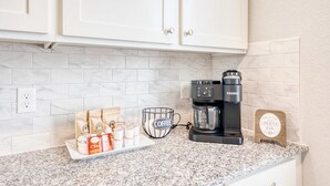 A kitchen with white cabinets, a coffee maker, a sign that says "Our Little Coffee Bar," and a tray with coffee cups and bags of coffee.