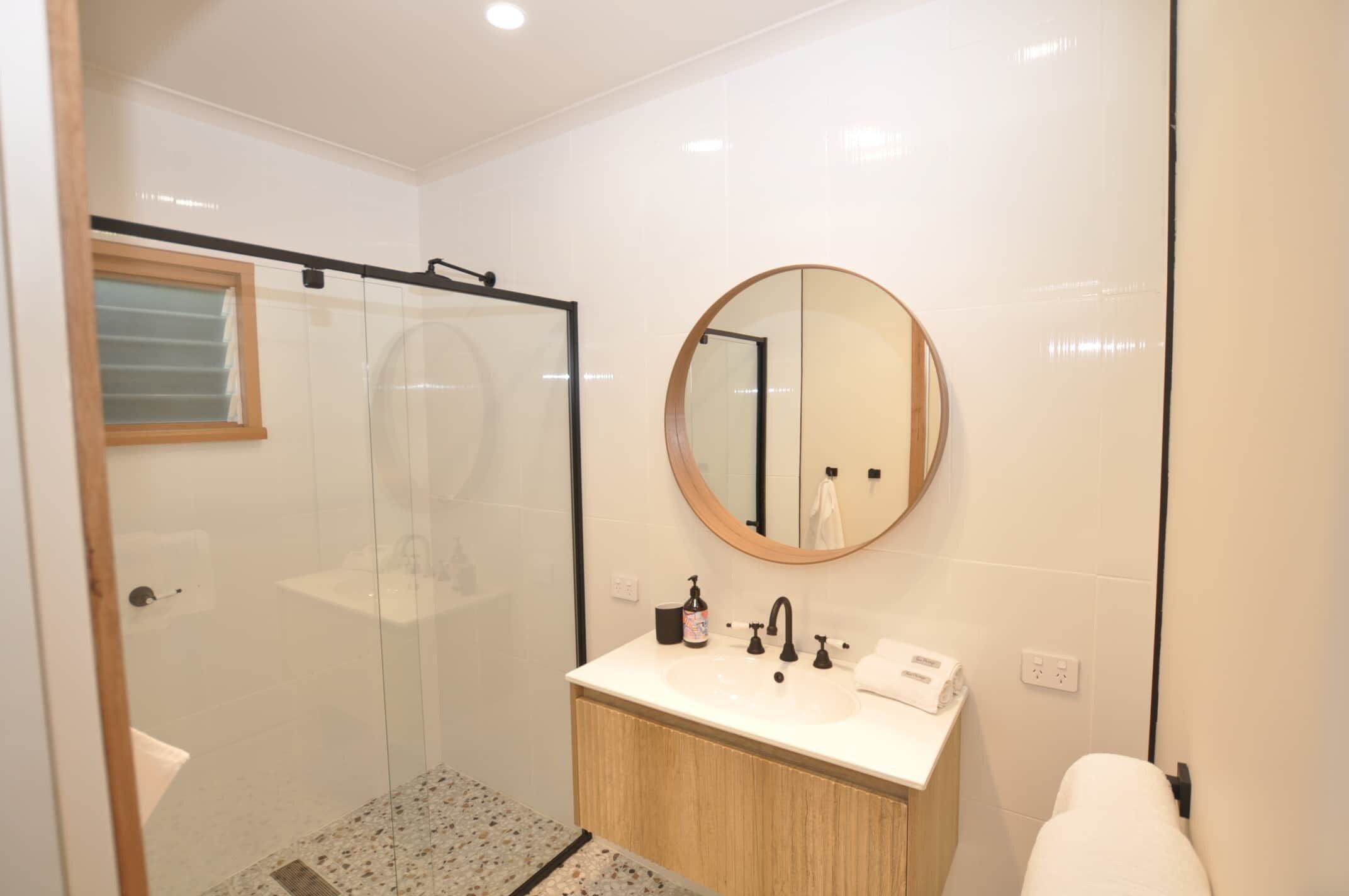 Un baño moderno con ducha envuelta en vidrio, un espejo redondo y un lavabo blanco con tocador de madera.