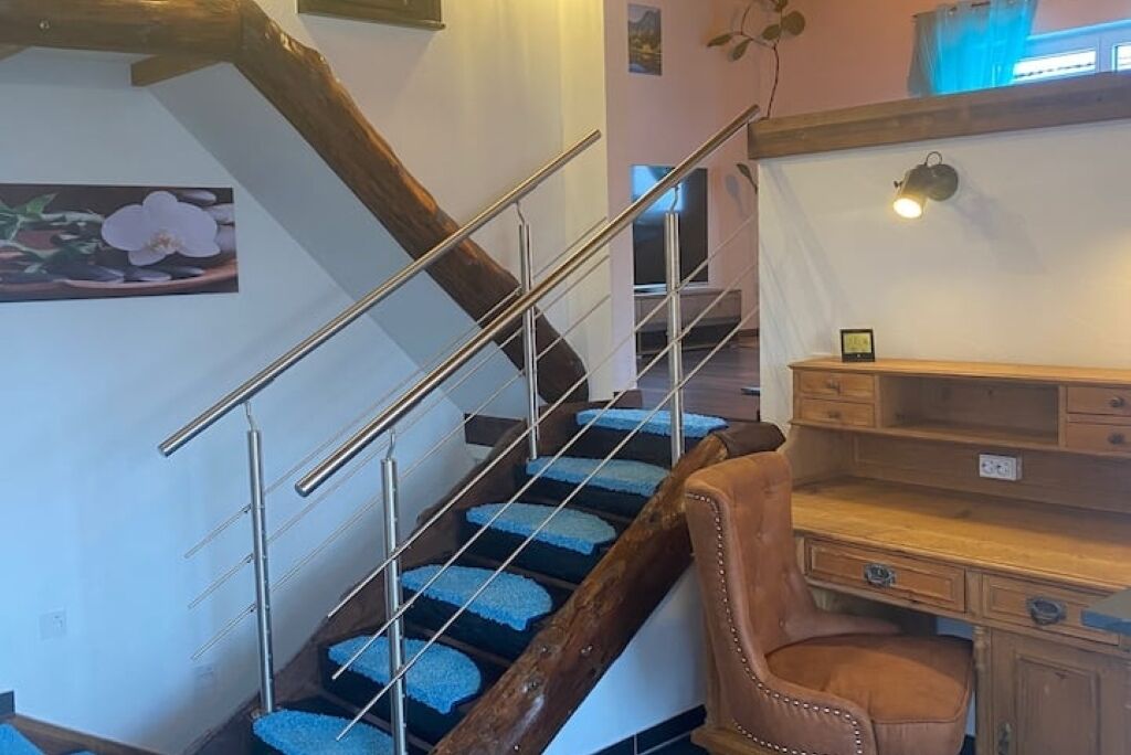 A staircase with a wooden handrail and blue carpeted steps, leading to a room with a wooden desk and a chair.