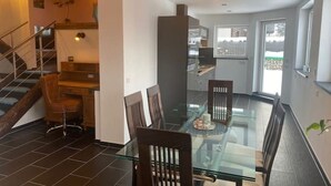 A modern dining area with a glass table and wooden chairs, a staircase with a wooden railing, and a view of the outside through a large window. - Cozy vacation home (Biessenhofen)