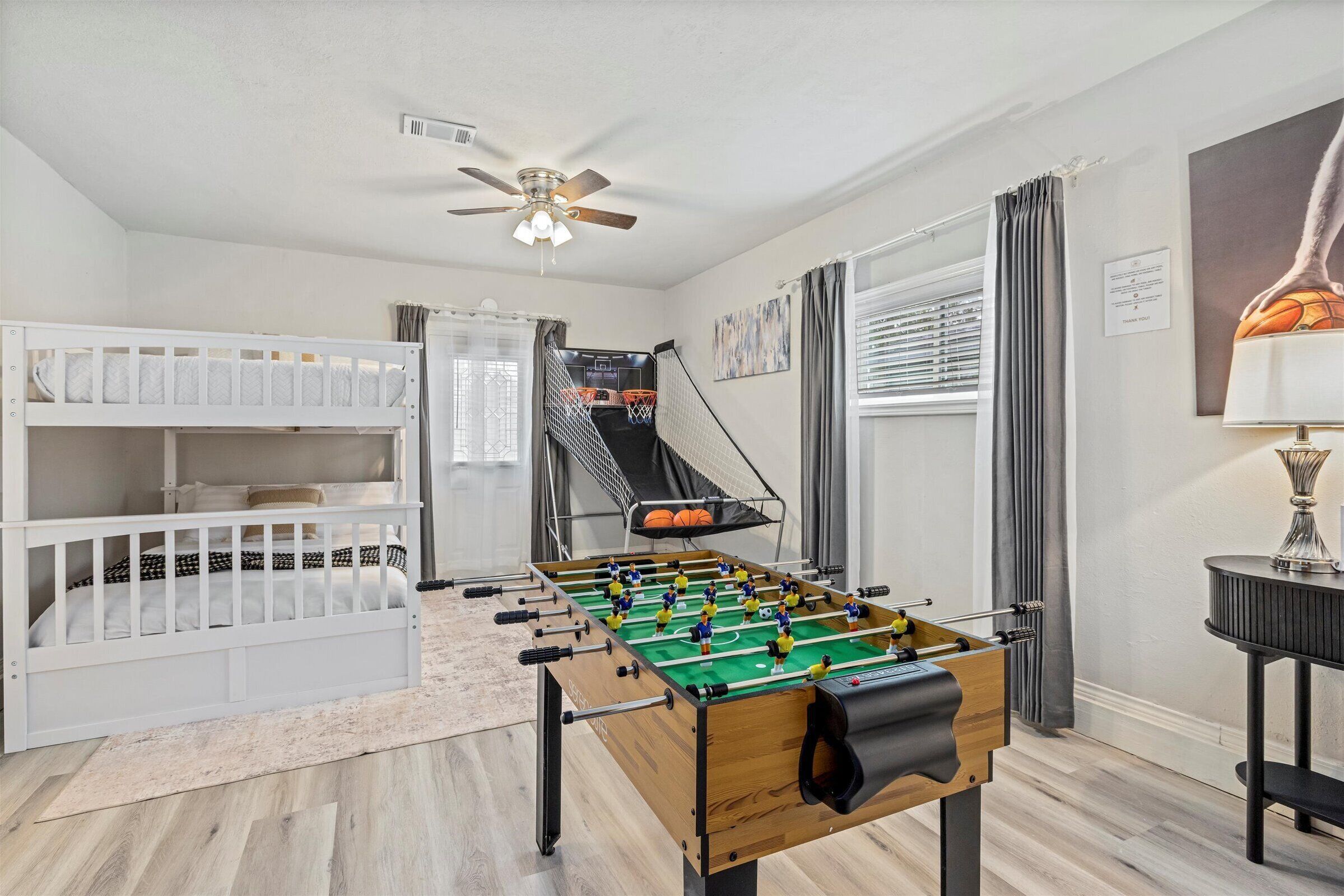 A room with a foosball table, bunk beds, a ceiling fan, and a window with curtains.