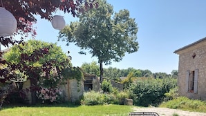 A rustic outdoor seating area with wicker chairs and a wooden table, surrounded by lush greenery and stone buildings.