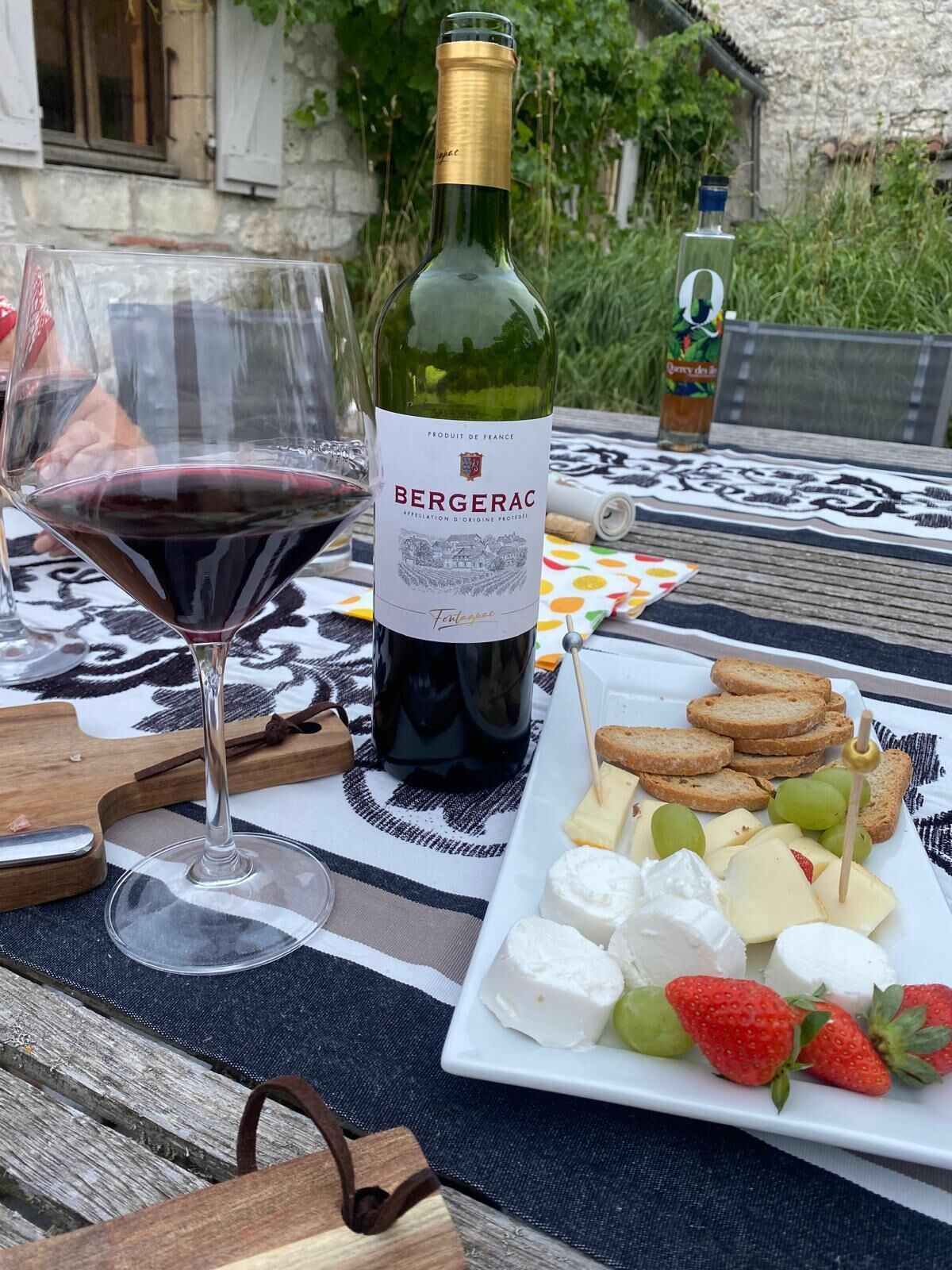 A bottle of Bergerac wine, a glass of red wine, and a cheese board with crackers and strawberries on an outdoor table.