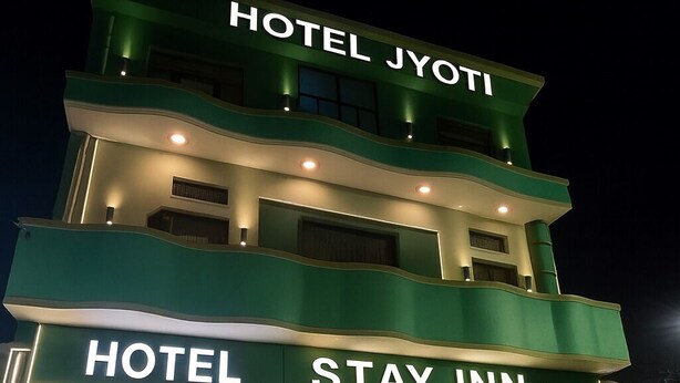 Un hôtel à plusieurs étages, nommé « Hotel Jyoti Stay Inn », doté d’un design moderne, avec une voiture garée devant et un chien près de l’entrée.