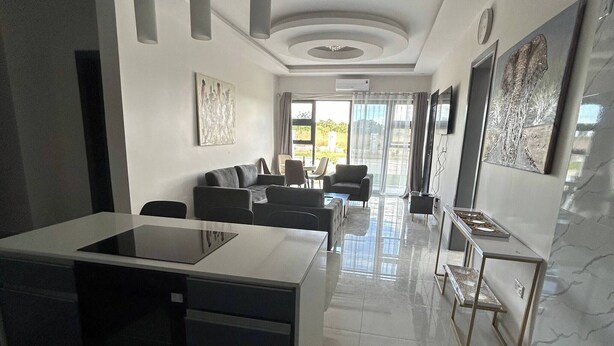 A modern kitchen with a central island, a dining area with a sofa and chairs, and a living room with a TV and artwork. - Luxurious Motherland Apartments (Lusaka)