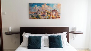 Una habitación de hotel con cama, dos almohadas, un cuadro de un paisaje urbano y una mesita de noche con lámpara.