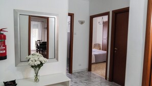 Un pasillo con pared blanca, un espejo, un jarrón con flores blancas, un extintor y una puerta que da a un dormitorio.