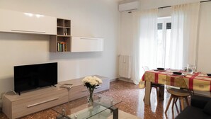 Una sala de estar moderna con un televisor de pantalla plana, una mesa de centro de vidrio, un mueble de madera y un comedor con una mesa preparada para dos personas.