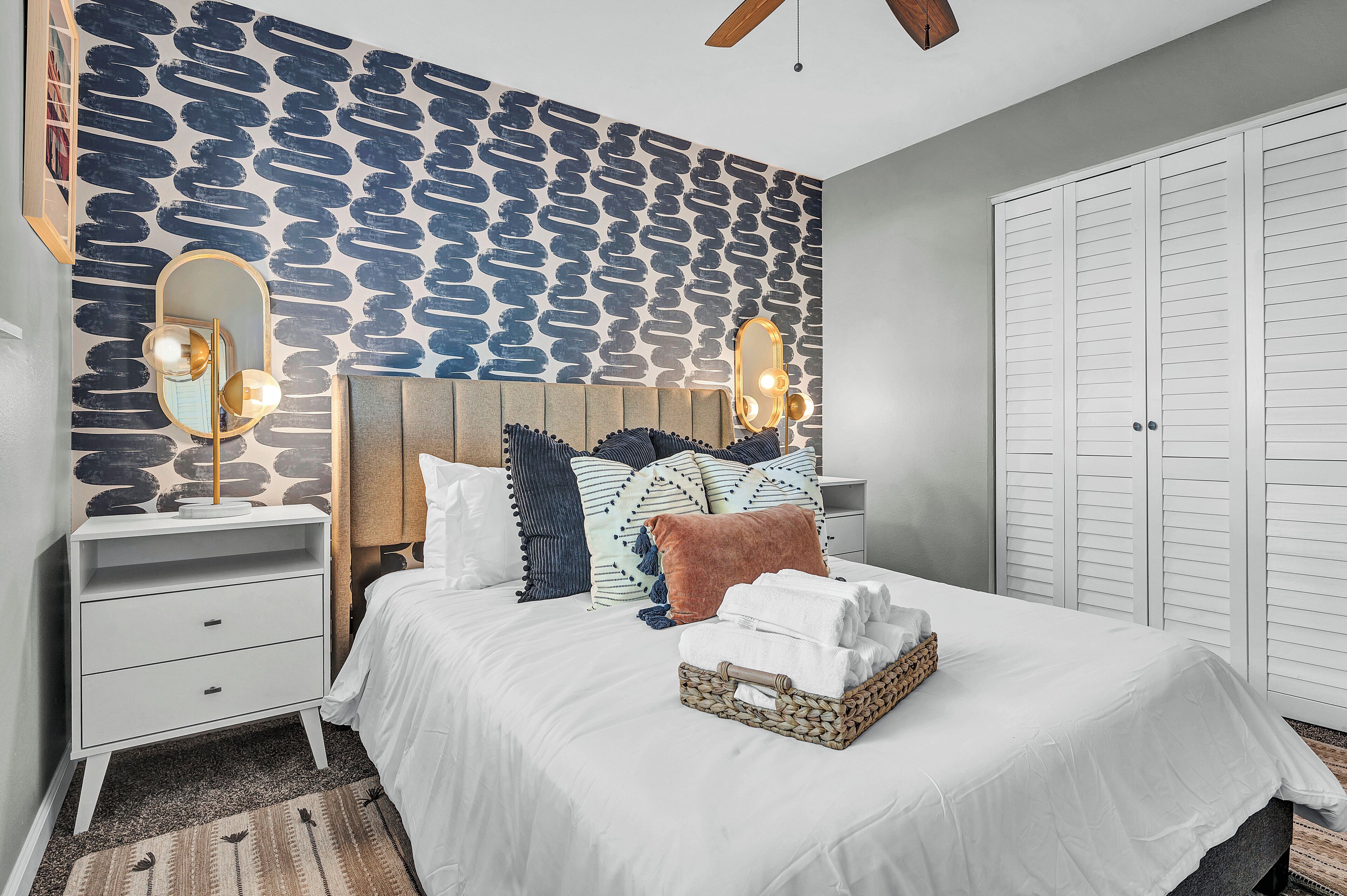 A neatly arranged bedroom with a patterned wall, a bed with white linens, a bedside table with a mirror, and a wardrobe.